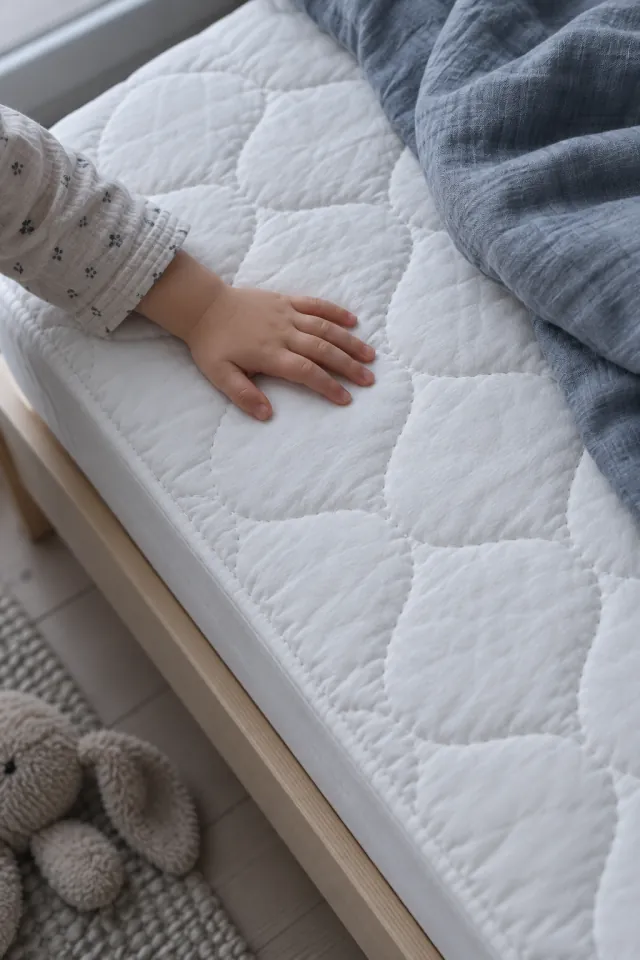 Mano de un niño sobre la funda acolchada blanca de un colchón infantil, con una manta gris azulada